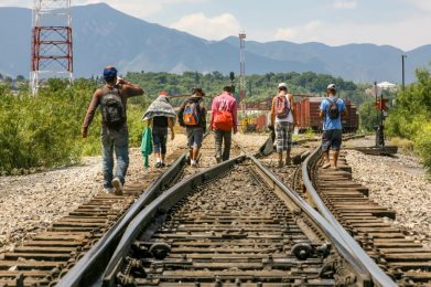 Group of people wearing backpacks and holding blankets, walking along railroad tracks
