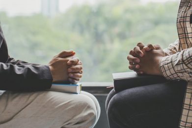 Two people sitting opposite one another with book of scripture on each of their laps, their hands folded