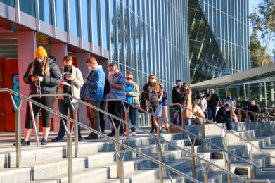 line of people wearing masks, standing outside a large glass building