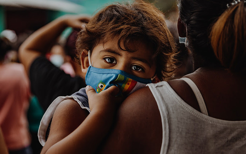 small child wearing mask, being carried in arms of mother